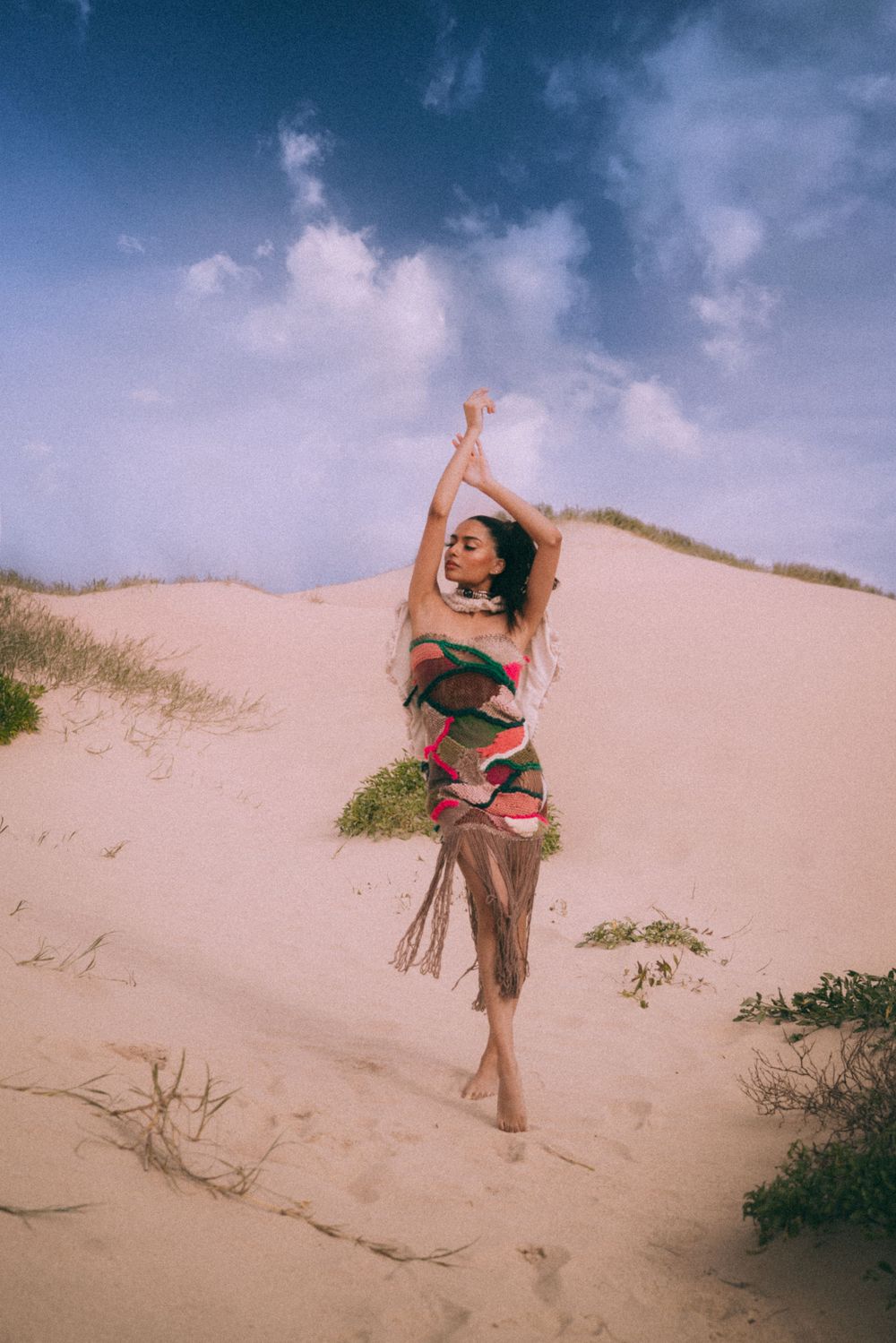 A person in a flowy patterned dress dances gracefully on sand dunes against a dreamy blue sky with white clouds.
