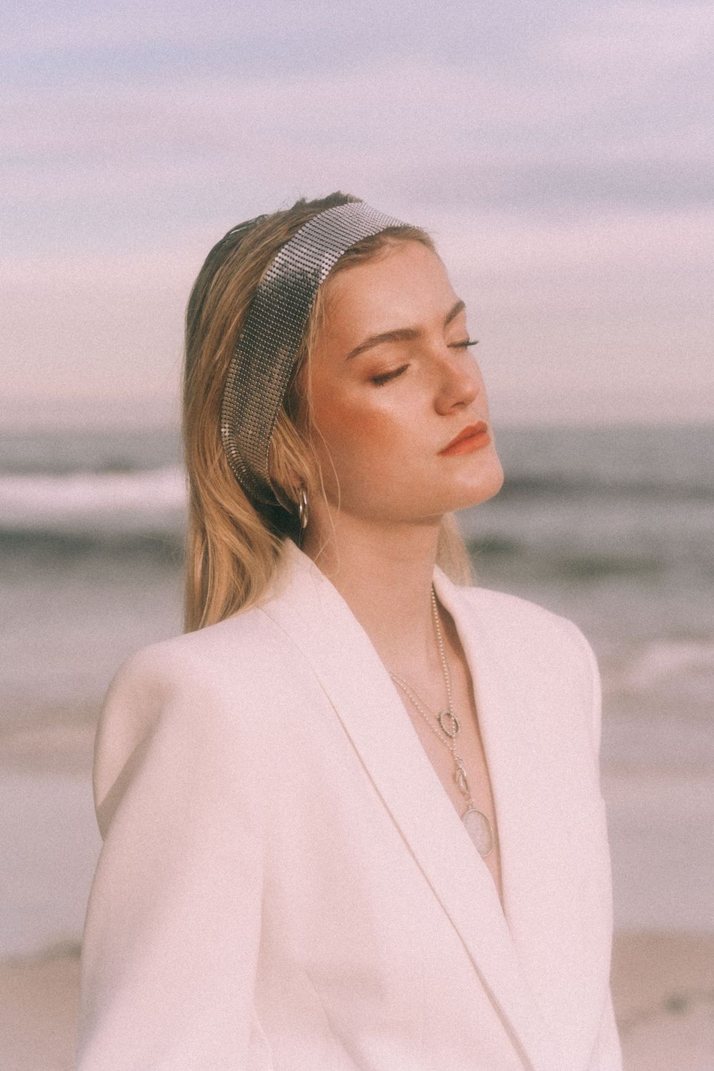 A serene portrait with a person wearing a white blazer and headband against a pink sunset beach background.