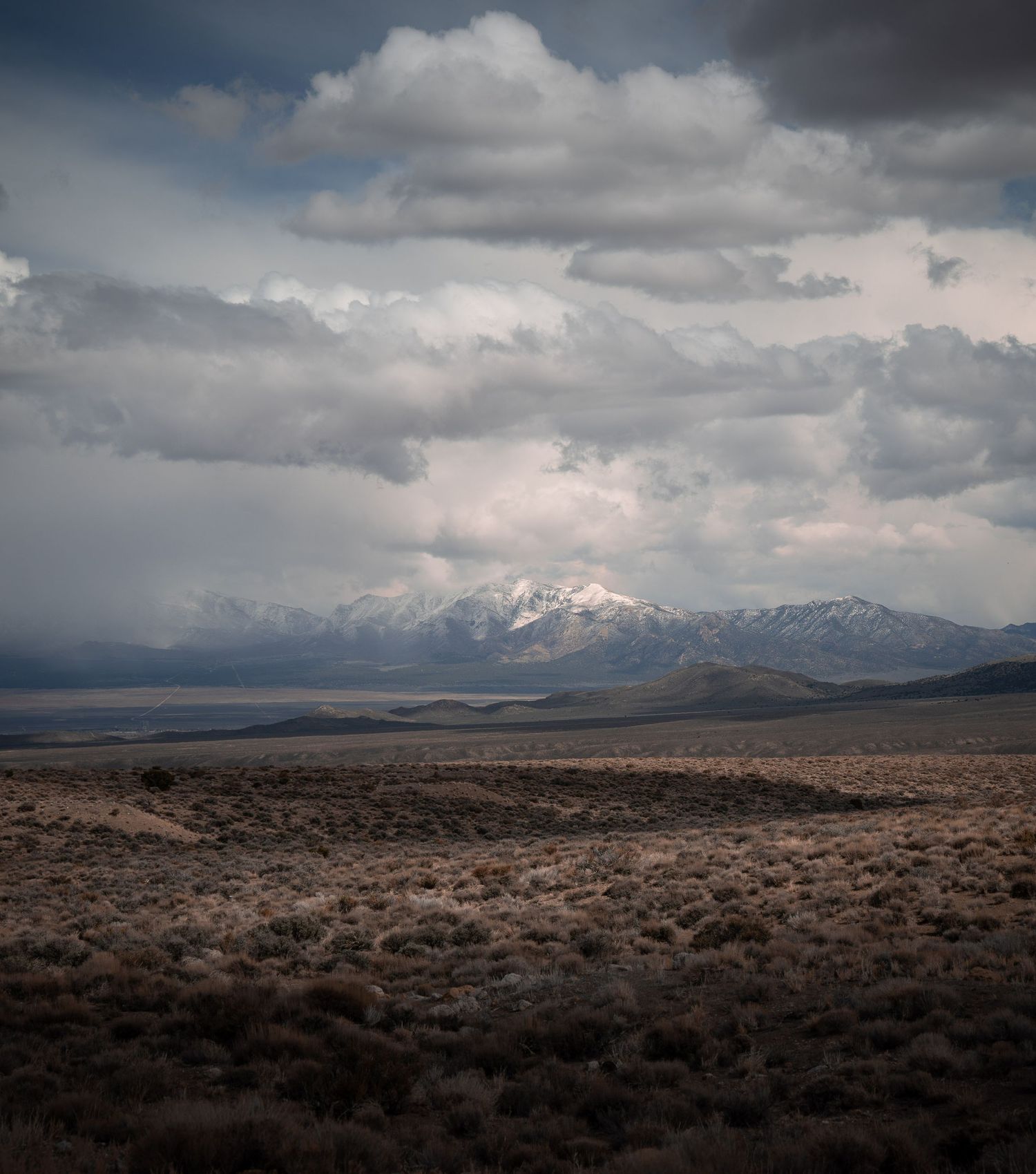 Dramatic stormy clouds roll over a rugged desert landscape with snow-capped mountains in the distance.
