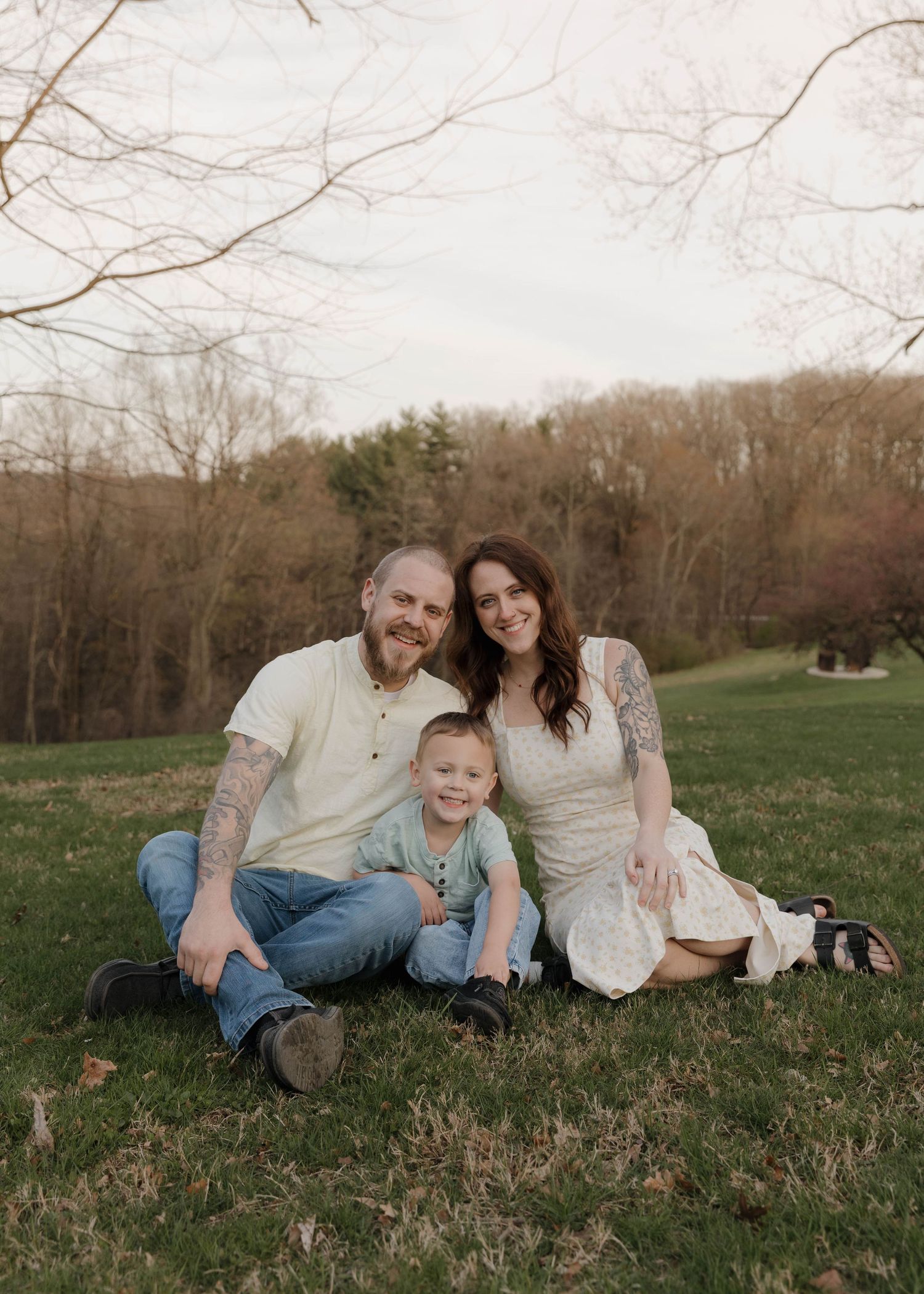 A family sits together on grass in a rustic outdoor setting during autumn with trees in the background.