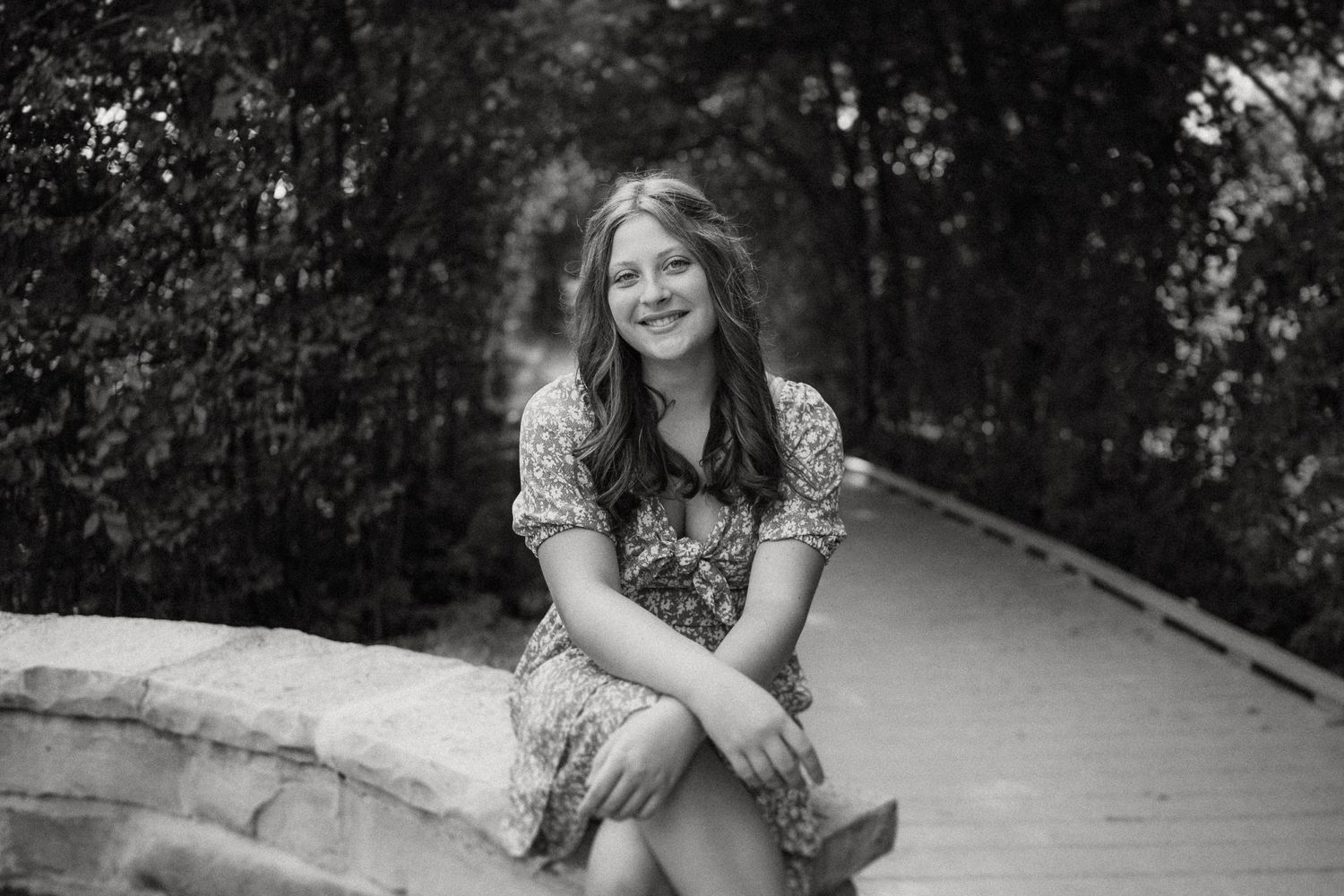 Black and white portrait of someone in a floral dress sitting outdoors on a curved wall in a wooded setting.