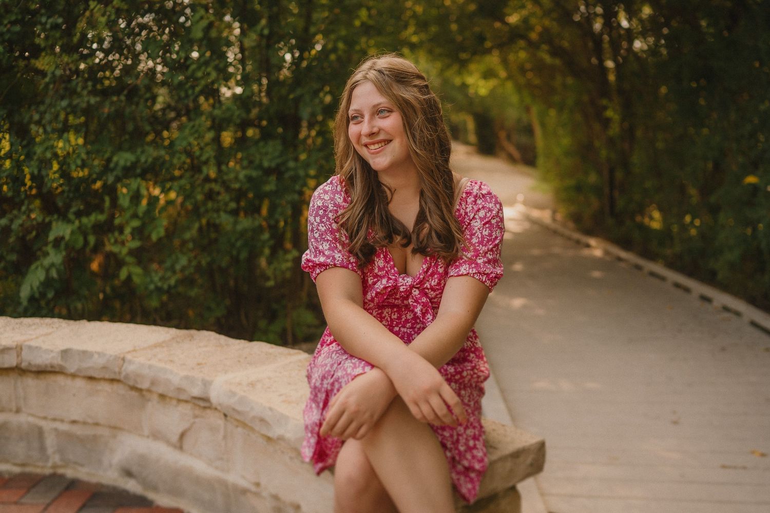 A person in a pink polka dot dress sits smiling on a stone wall along a tree-lined path on a sunny day.