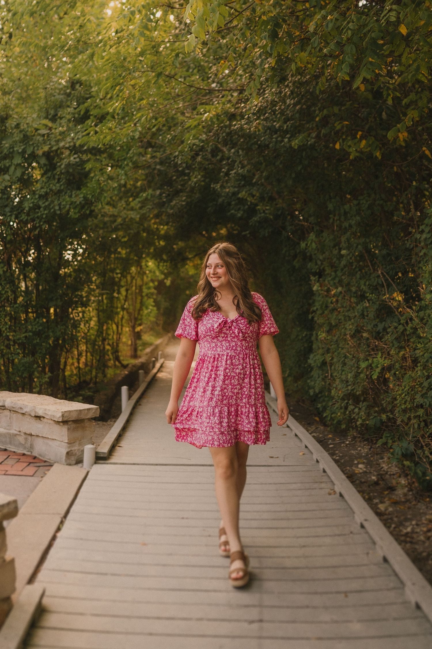 Person in a flowing pink summer dress walks along a wooden boardwalk path surrounded by lush green foliage.