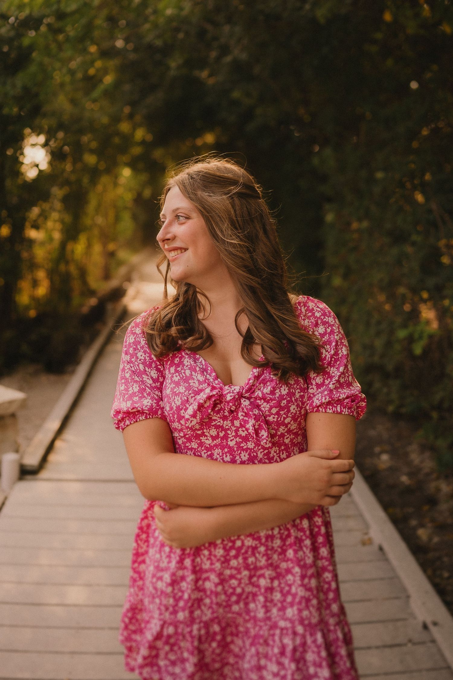 A person in a pink floral dress stands on a wooden boardwalk path surrounded by autumn foliage at sunset.