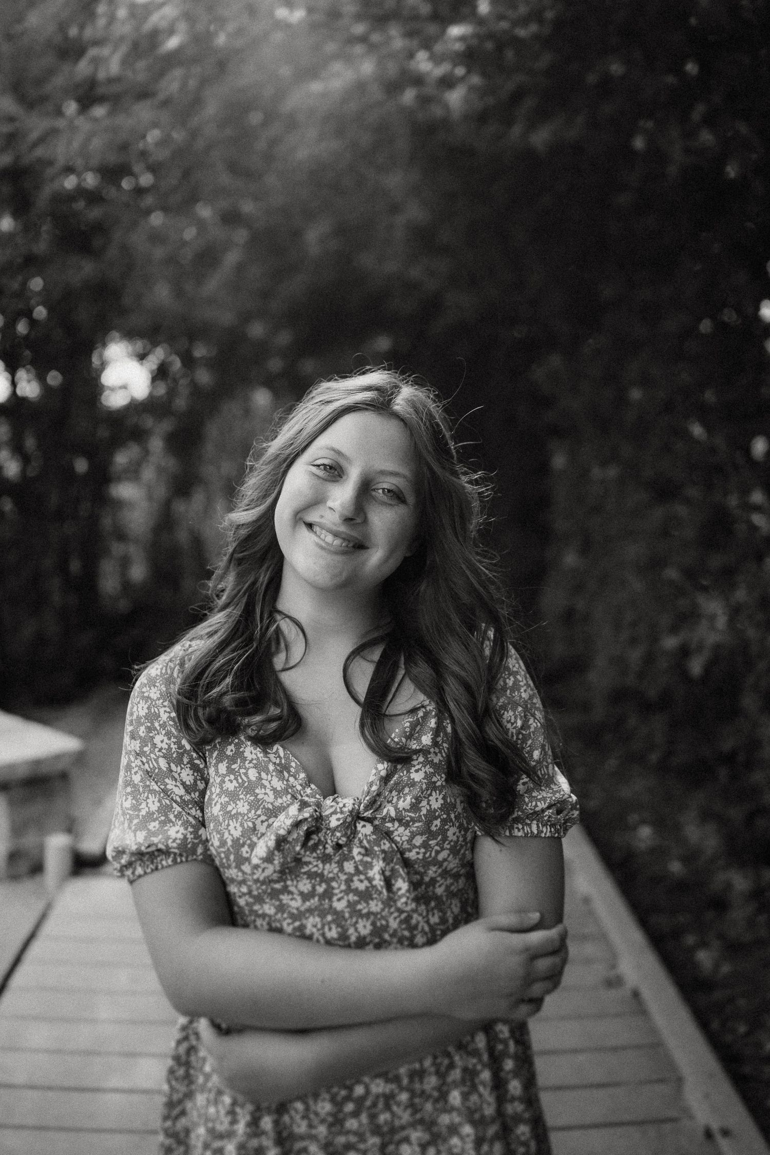 Black and white portrait of a person in a floral dress smiling while standing on a wooden walkway outdoors.