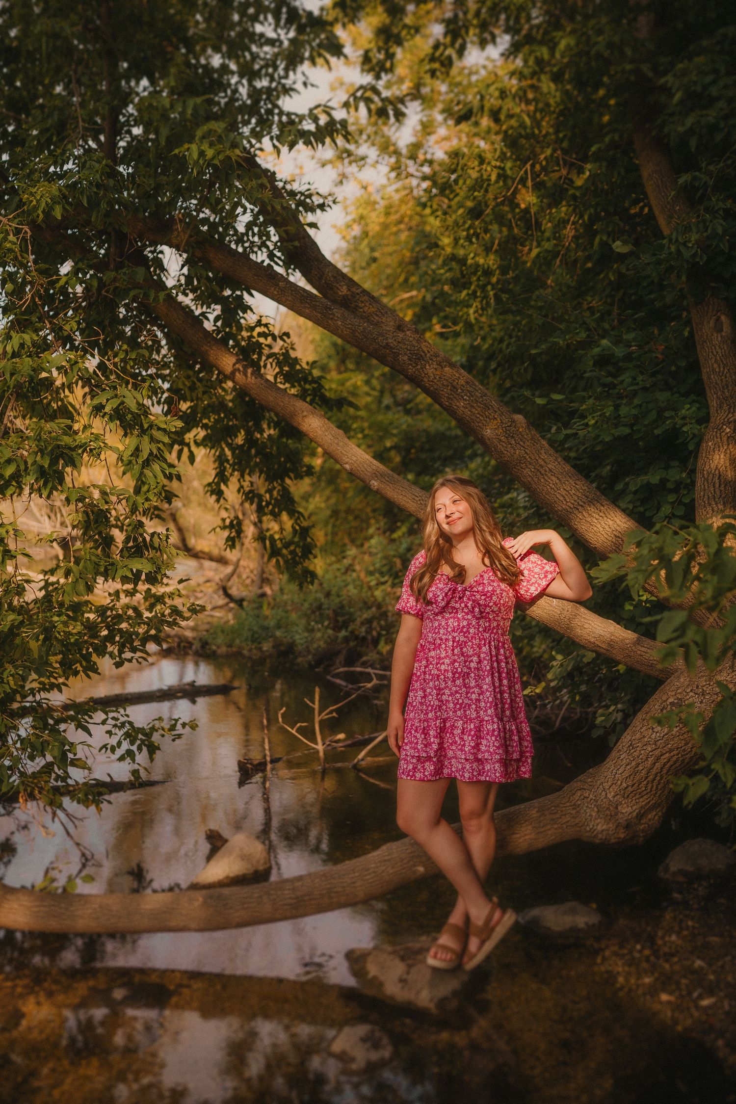 A person in a pink floral dress poses by a tranquil pond with overhanging trees during golden hour.