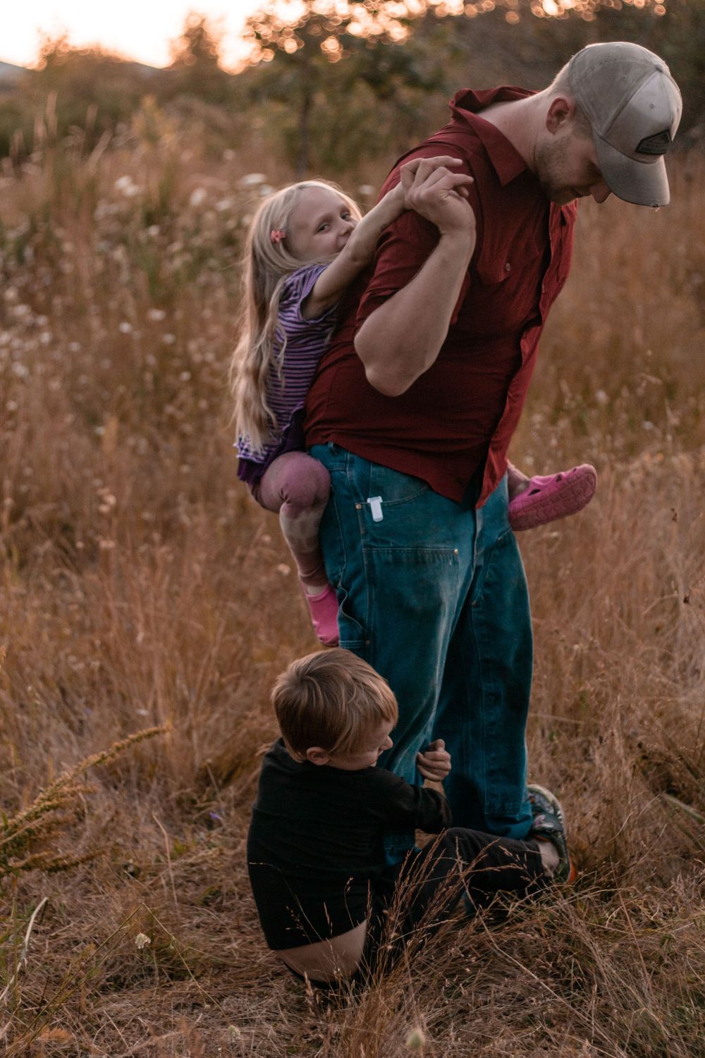 Father holding young blonde girl while toddler boy clings to his leg in golden field at sunset.