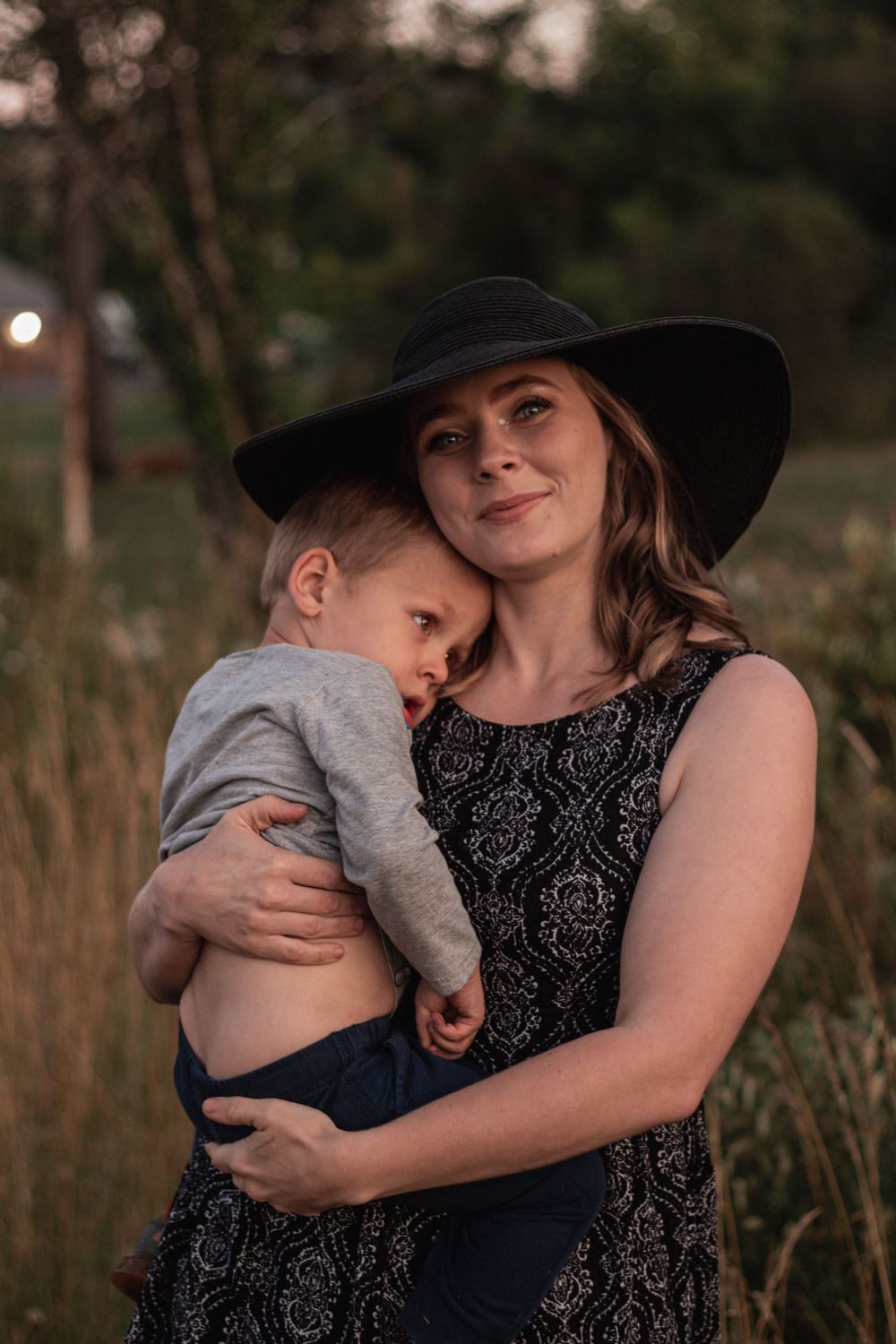 A woman in a wide-brim hat tenderly holds a sleepy toddler in an outdoor setting.