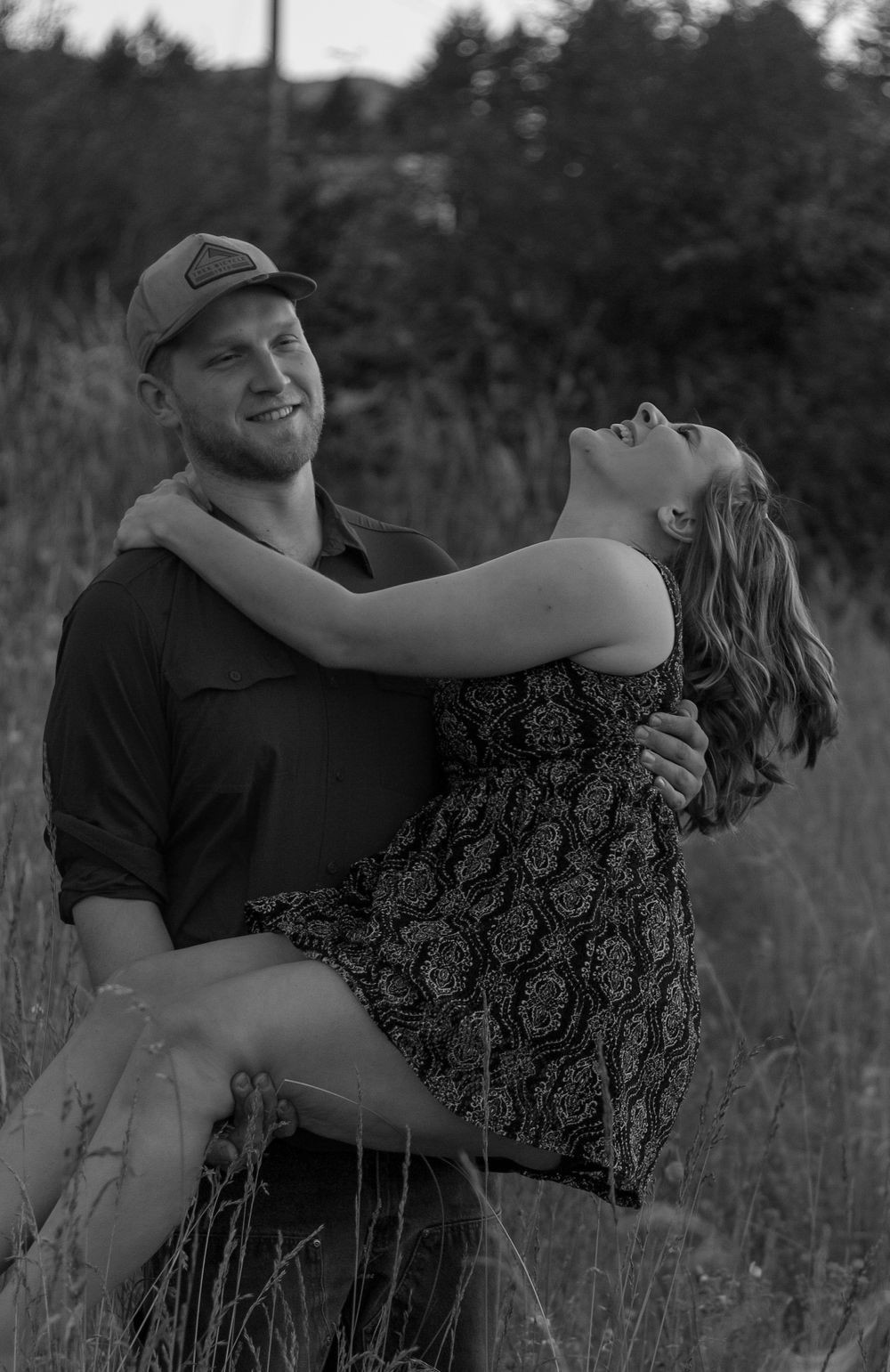 Black and white romantic photo of a couple in a field, woman leaning back joyfully.