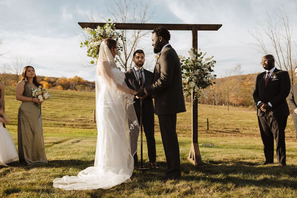 An outdoor autumn wedding ceremony takes place under a wooden arch with rolling hills in the background in sussex county nj.