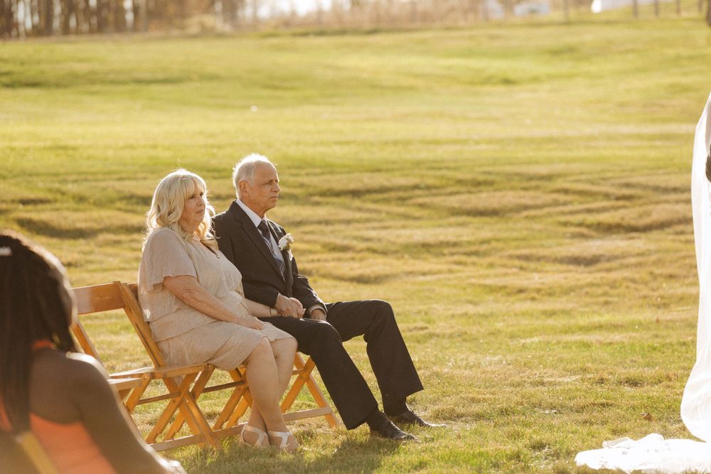 Two people sit on wooden chairs in a sunlit grassy field during an outdoor gathering at golden hour near vernon nj.