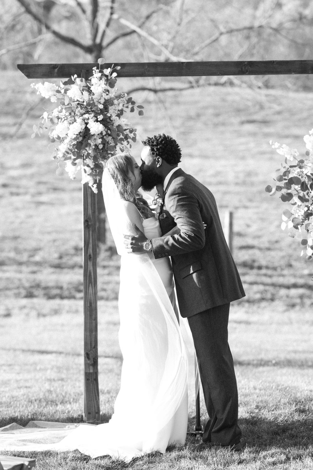 A black and white photo of a couple sharing a kiss under a wooden arch decorated with floral arrangements outdoors near vernon nj.