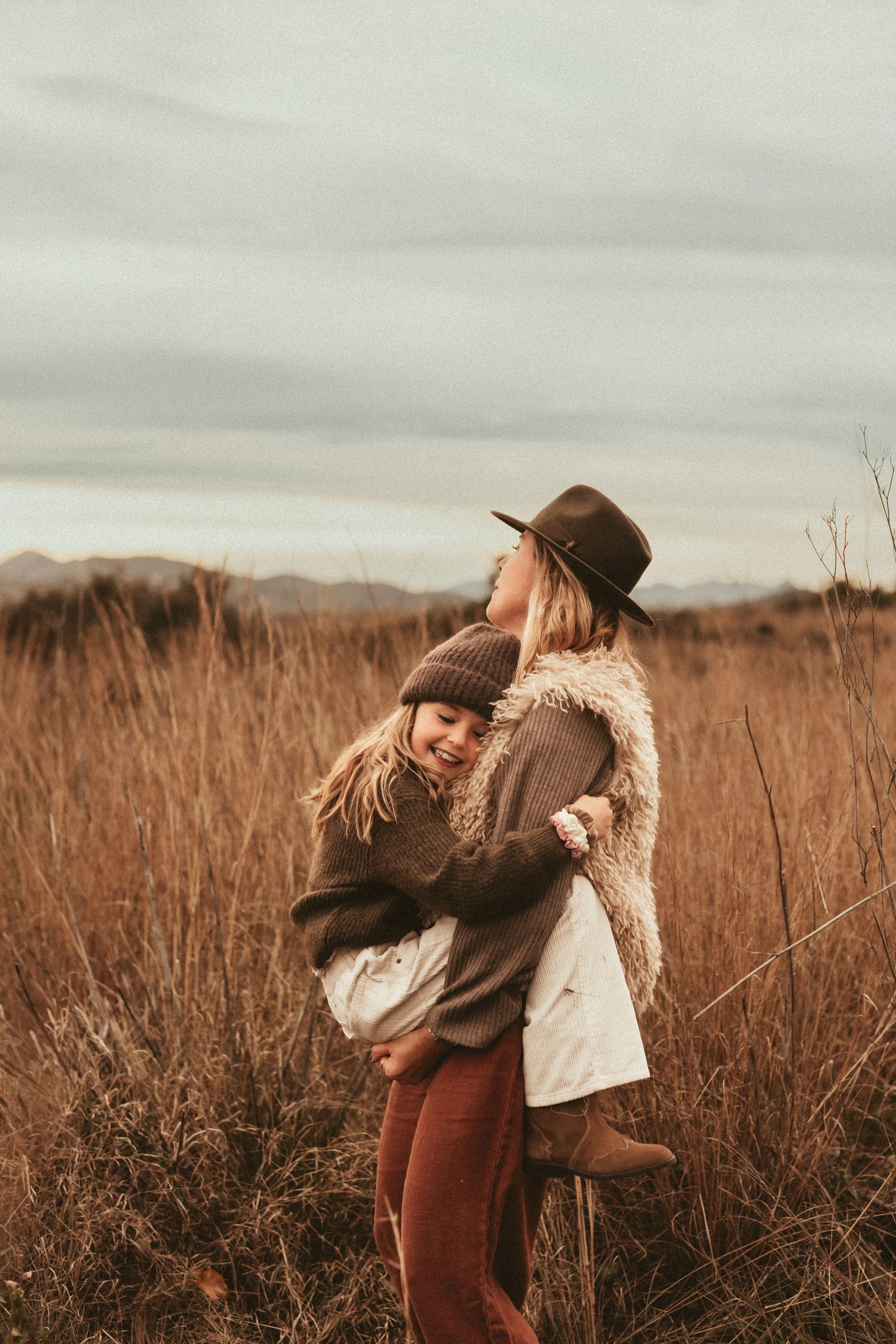 Mother and daughter  share a joyful moment together outdoors during golden hour in a field of tall grass, with Denia Photographer
