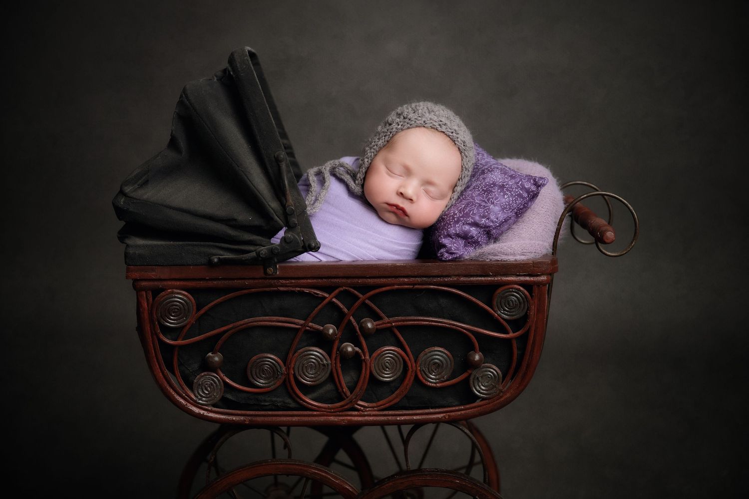 A sleeping baby nestled in a vintage wooden carriage with purple and black cushions against a dark background.