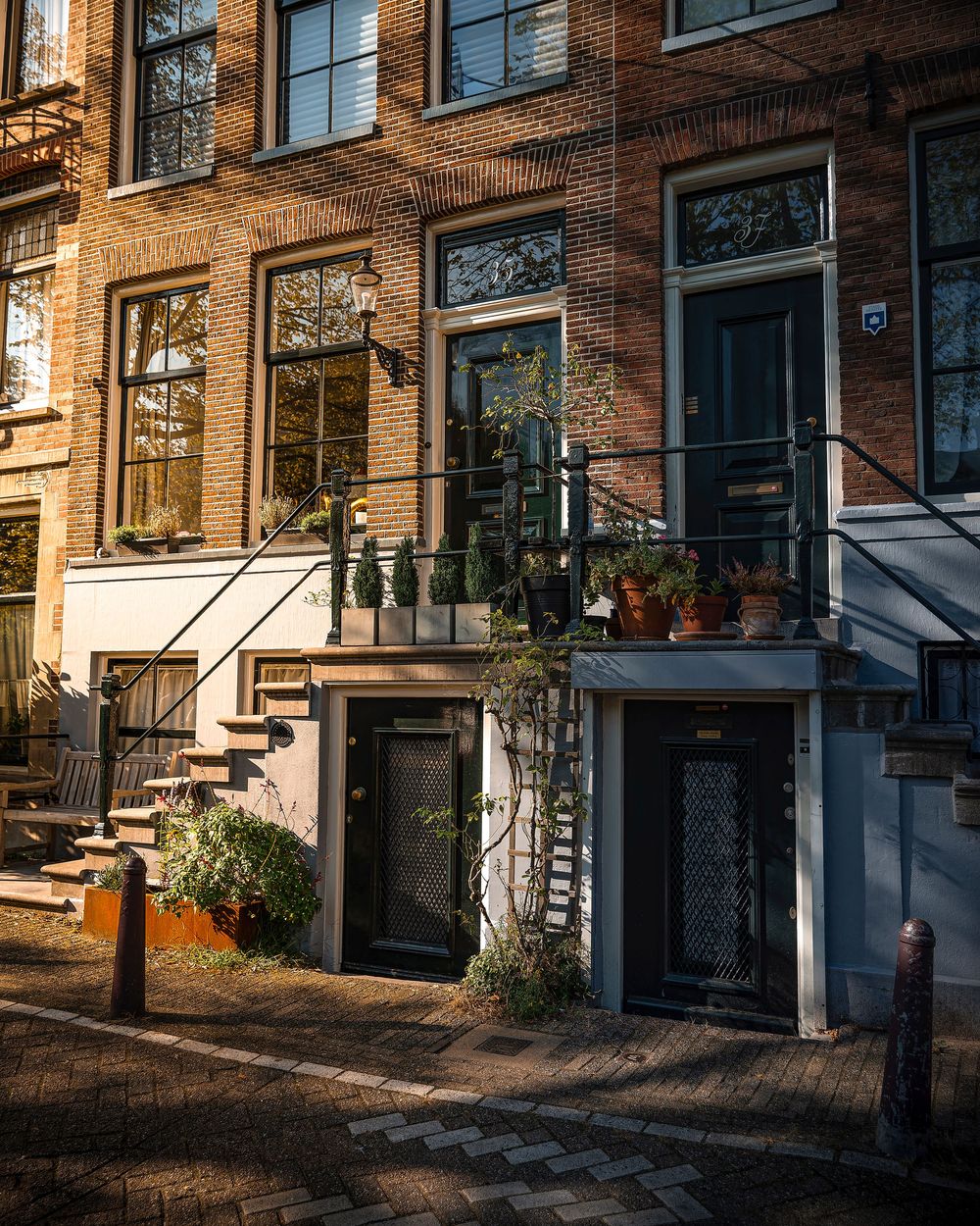 Historic Amsterdam canal house with brick facade, blue doors, iron staircases, and potted plants in warm golden sunlight.