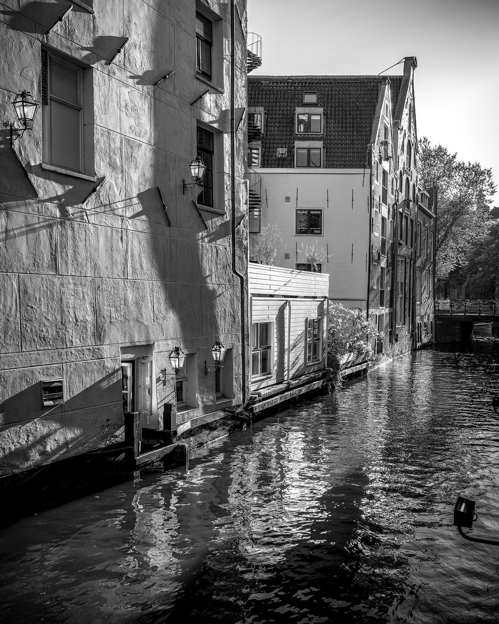 Black and white photo of a narrow Amsterdam canal flanked by historic buildings reflecting in dark water.