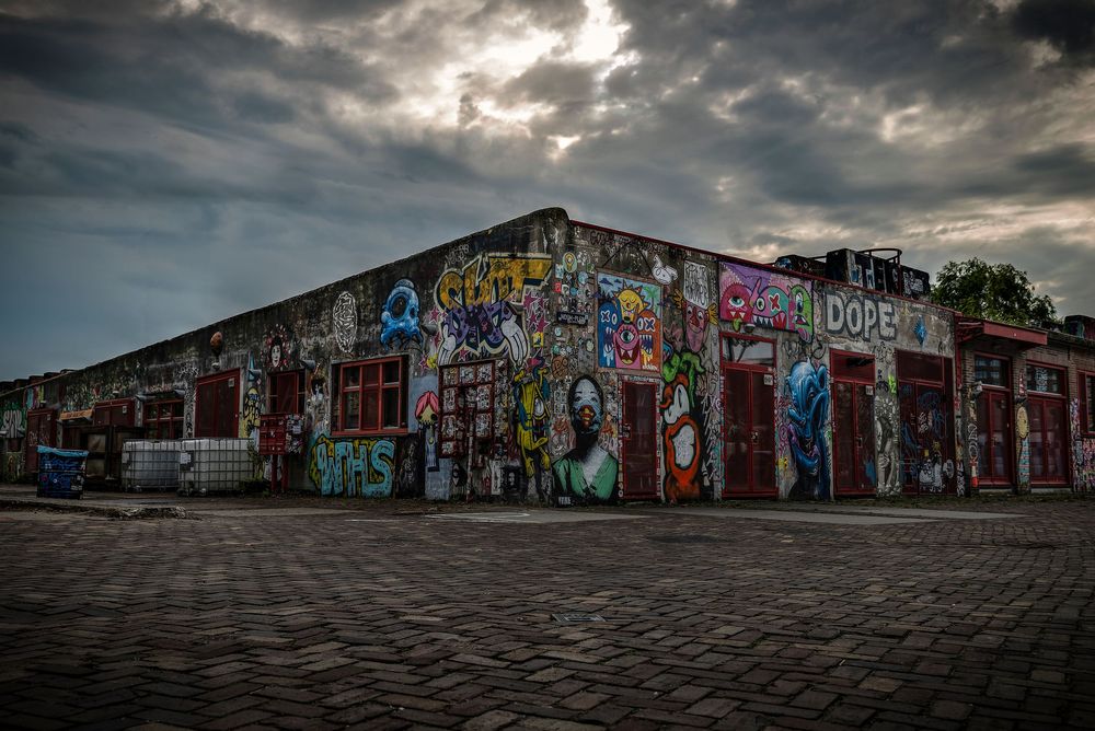 Colorful graffiti murals cover a brick building under dramatic cloudy skies on a cobblestone street.