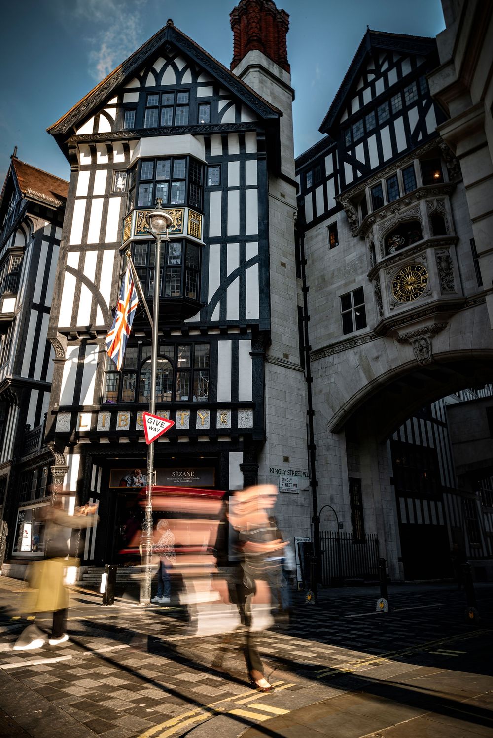 Motion-blurred figures walking past Tudor-style buildings with dramatic shadows in an historic English town street.