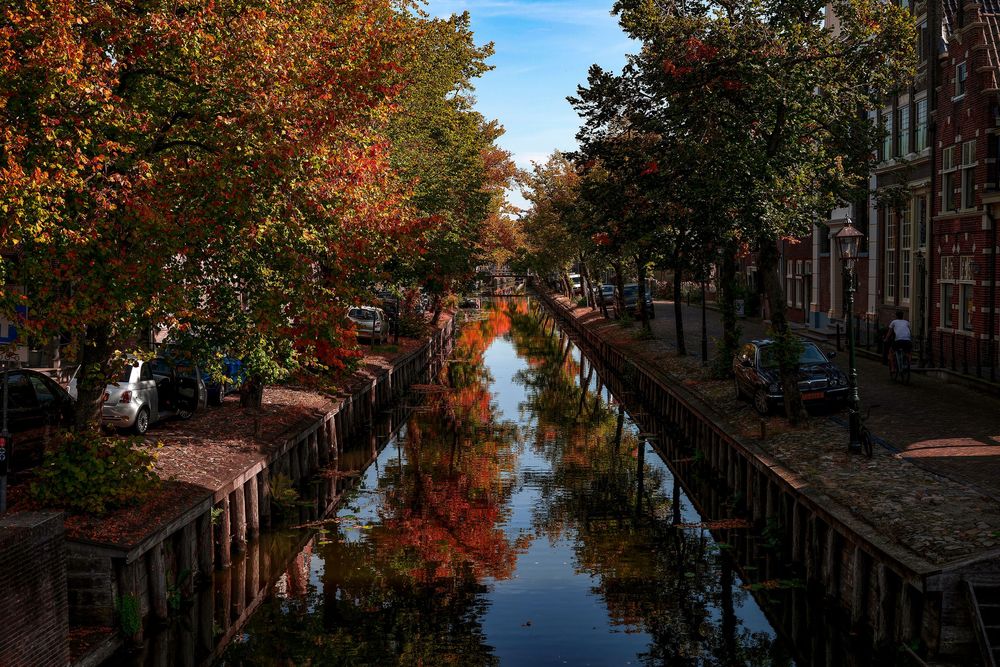 Autumn canal in Amsterdam with colorful red and orange trees reflecting in calm water, flanked by historic brick buildings.