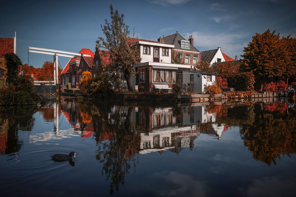 Autumn houses reflecting in calm canal water with colorful trees and blue sky in a Dutch village setting.