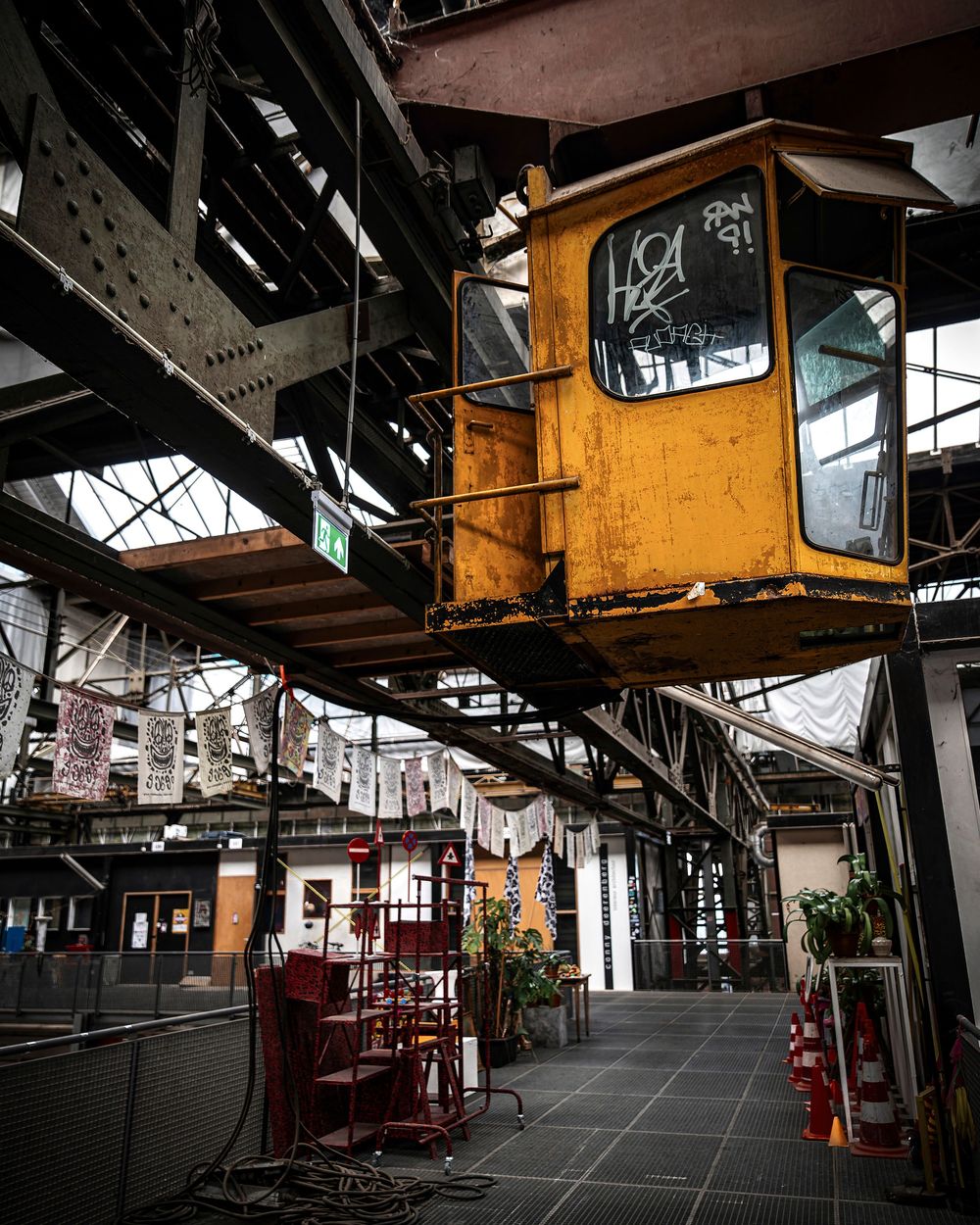Abandoned yellow crane cab suspended inside a gritty industrial warehouse with graffiti and red chairs below.