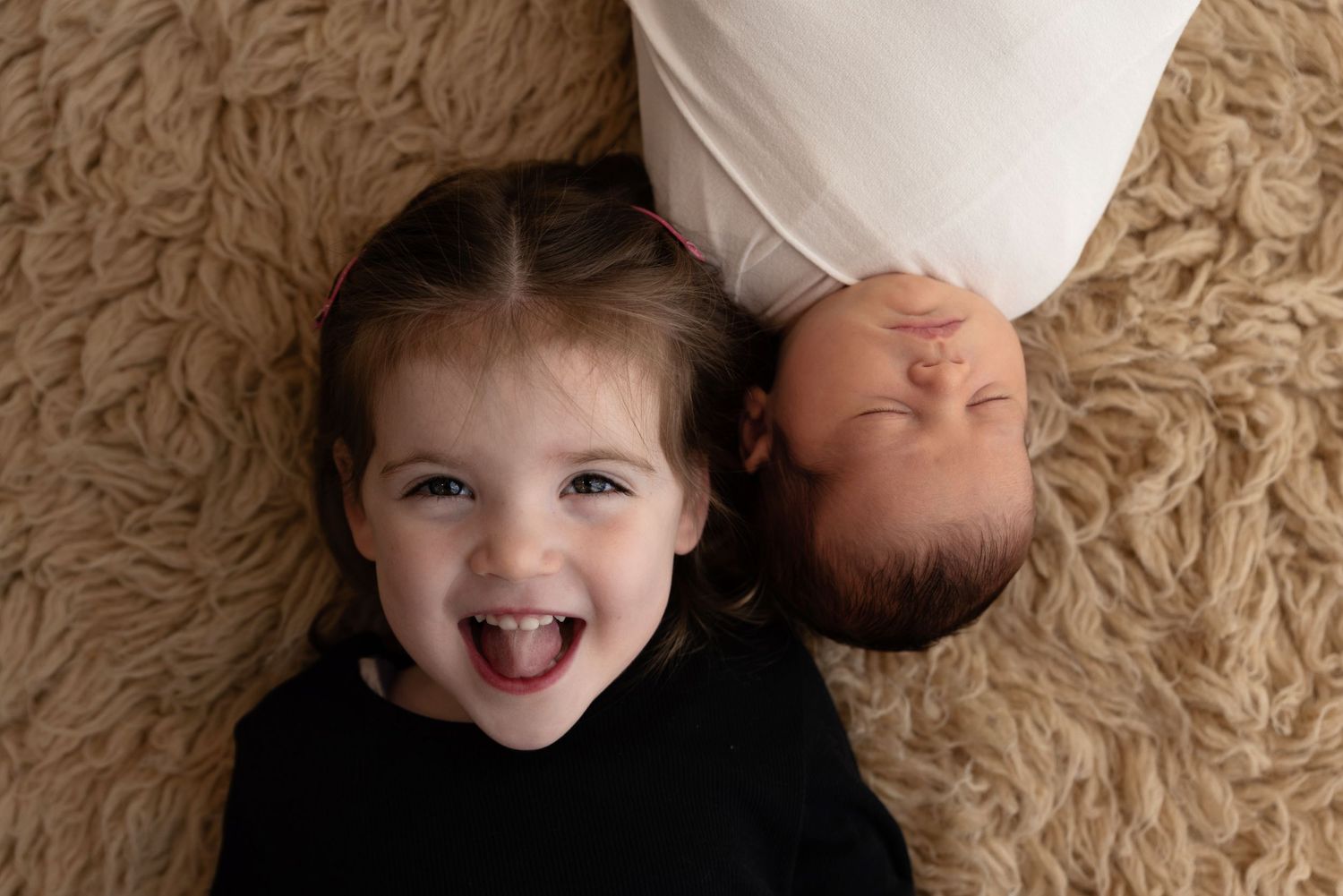 Two children lying on a beige shag carpet, one making a playful expression while wearing black.