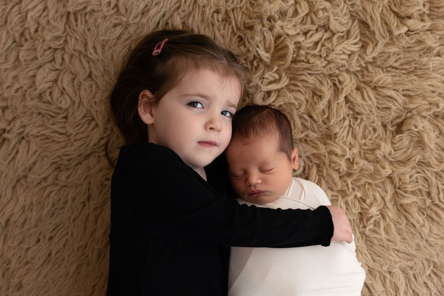 A young child in black hugging a baby wrapped in white on a textured beige carpet.