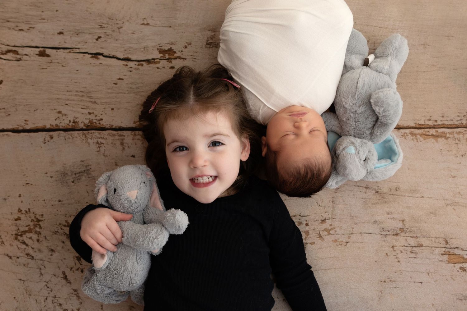 Two children lying on a rustic wooden floor with matching gray stuffed elephant toys.
