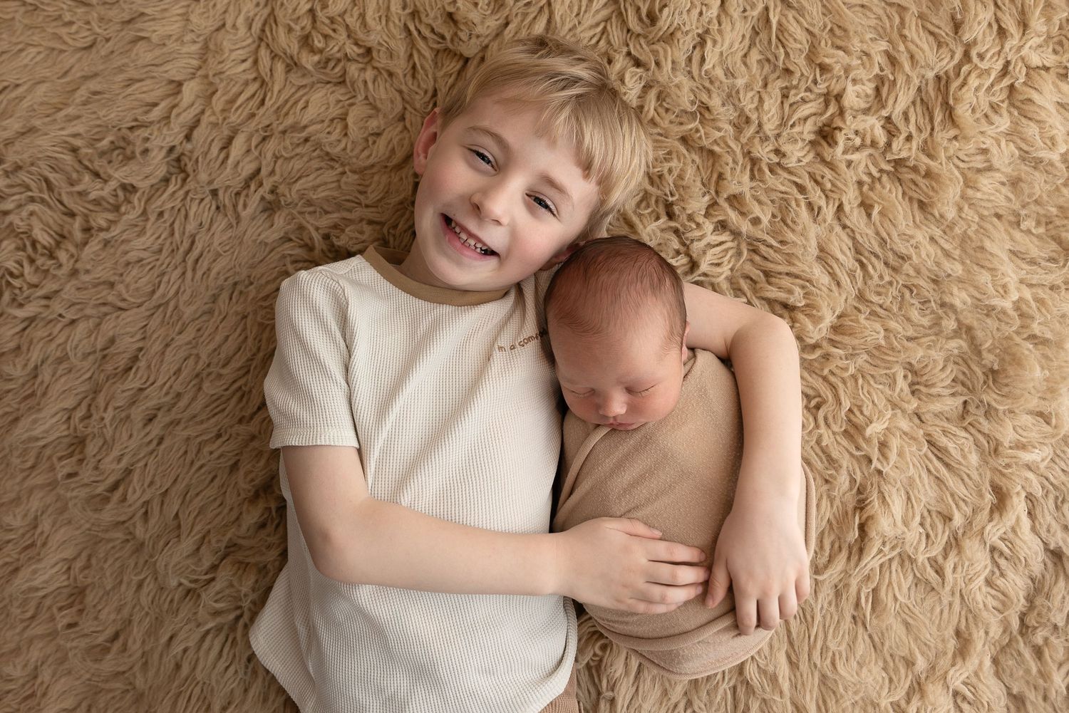 Two siblings lying together on plush shag carpet share tender moment during newborn photoshoot.