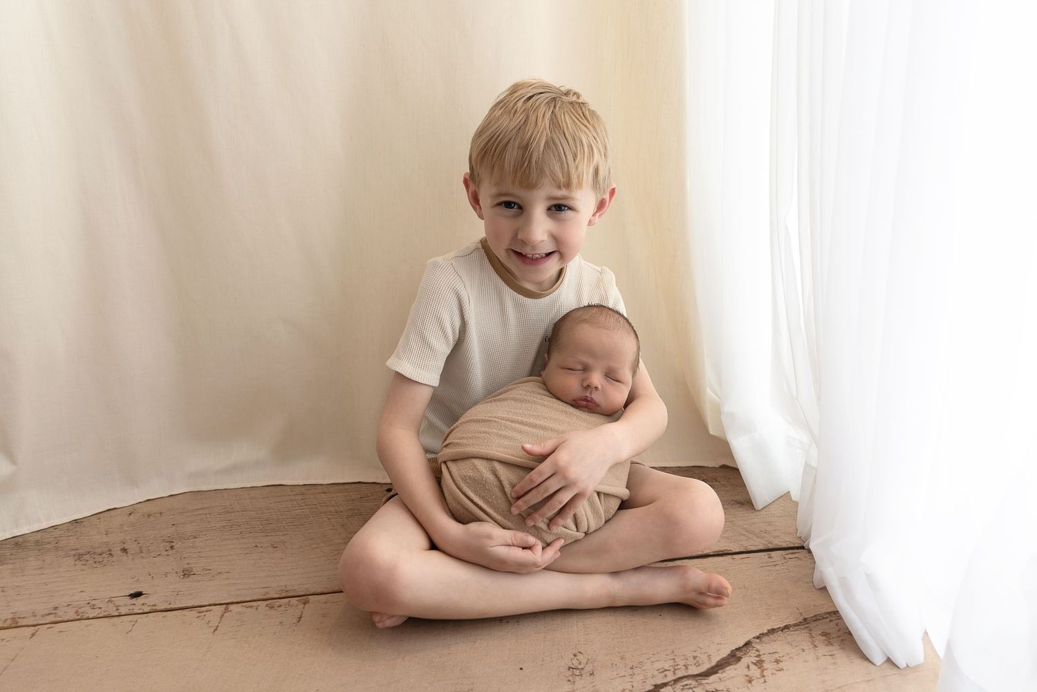 Two children sit together on a brown floor against a white curtained wall, showcasing a sweet sibling moment.