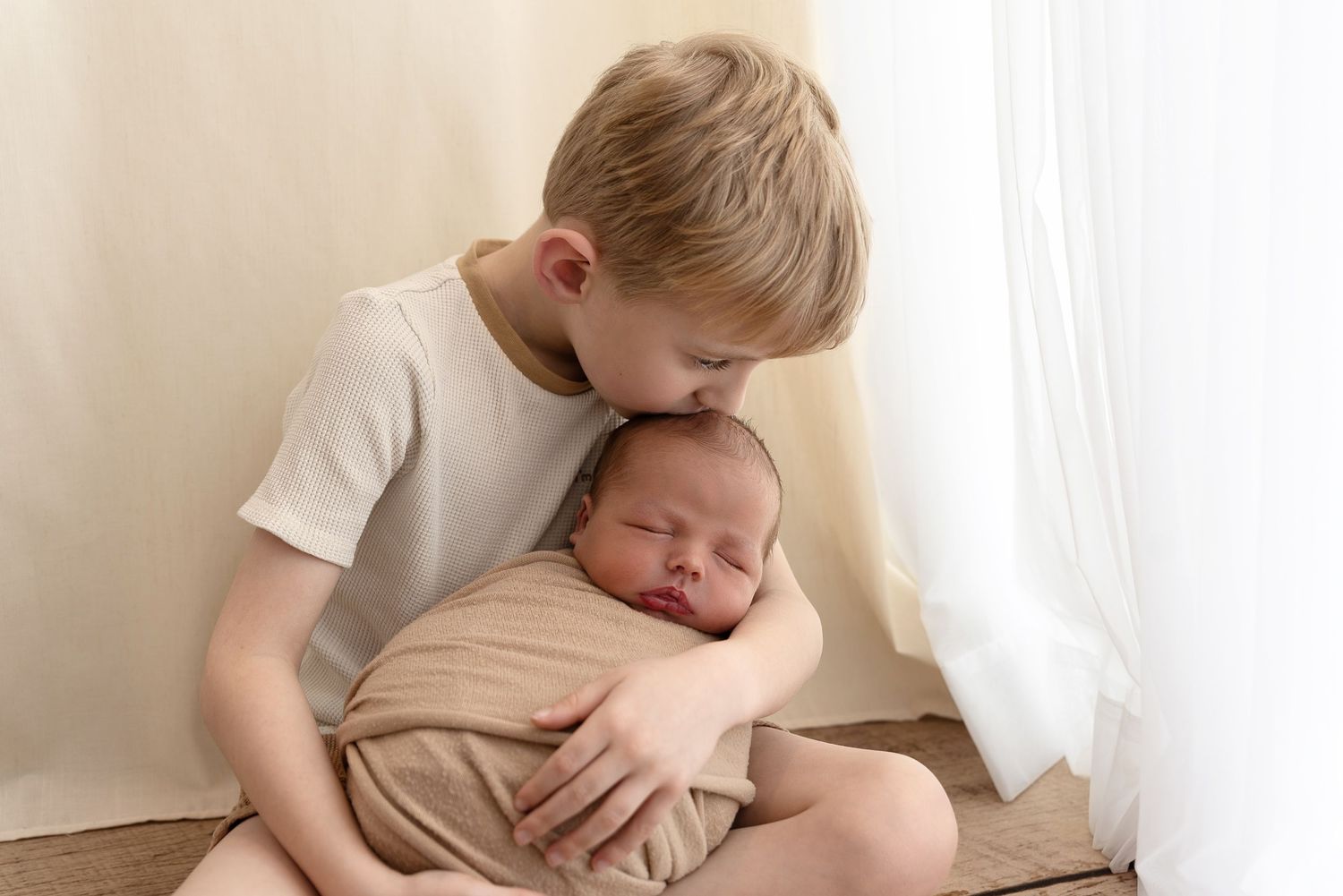 Young child in a white shirt sweetly embraces a swaddled newborn baby against a light background.
