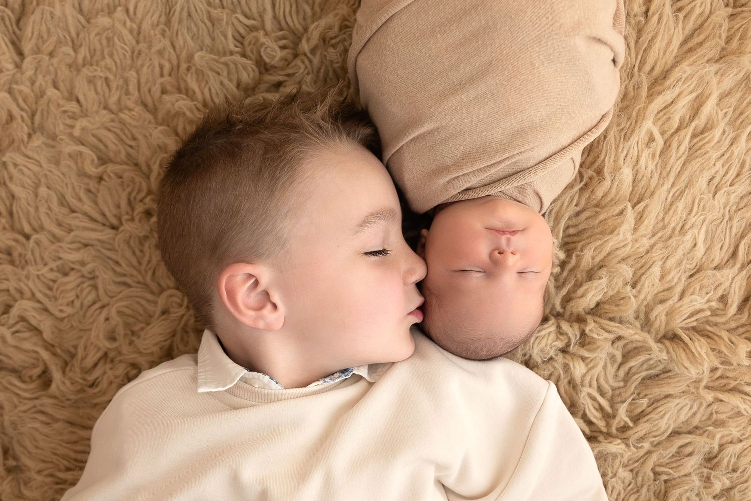 Two sleeping babies wearing white clothing lie face to face on a beige plush rug.