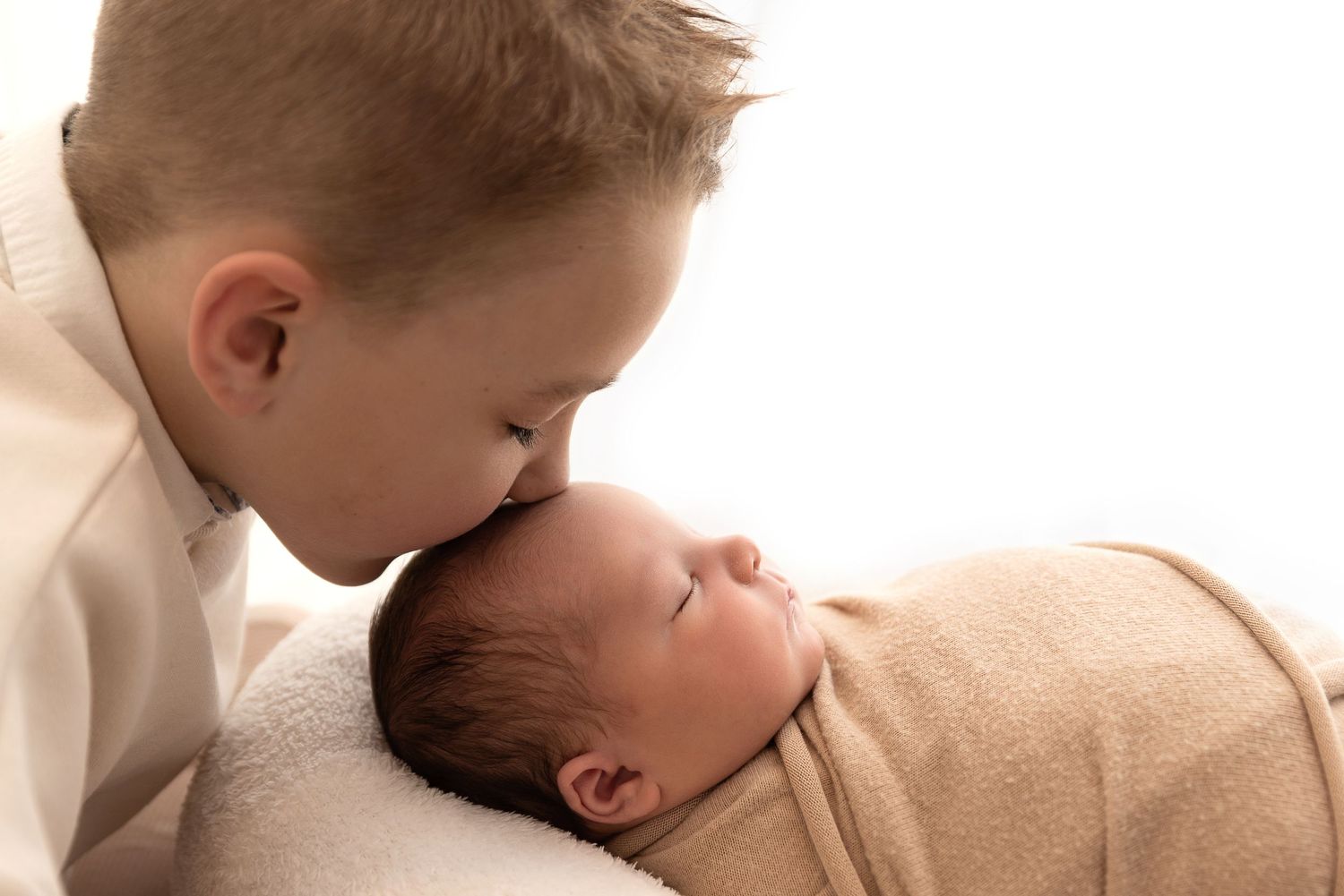 Two children close together with one gently leaning over to look at the other who is swaddled in a tan blanket.