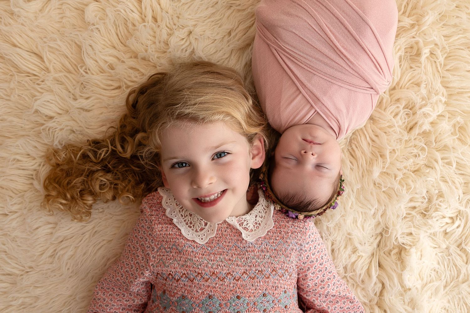 Two people wearing pink sweaters lie on a beige carpet while smiling at the camera.
