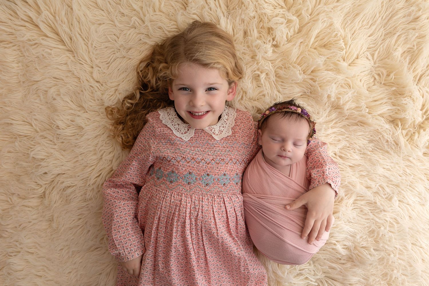 Two siblings pose together on beige carpet, older wearing pink smocked dress during professional photoshoot.