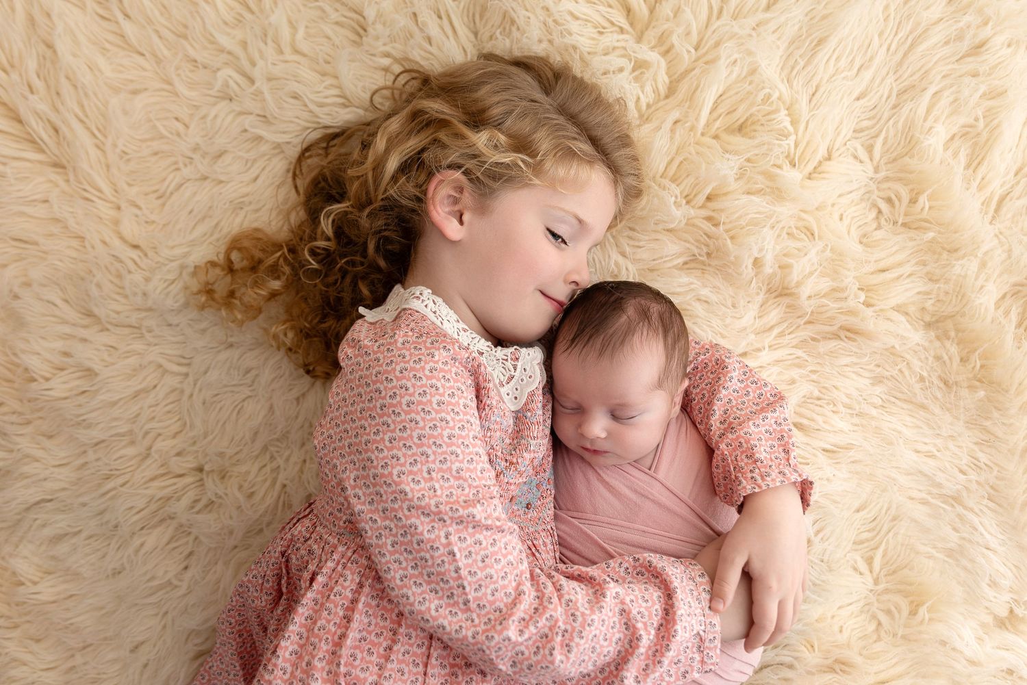 Two figures in soft pink clothing lie together on a cream-colored fluffy rug in a tender pose together.