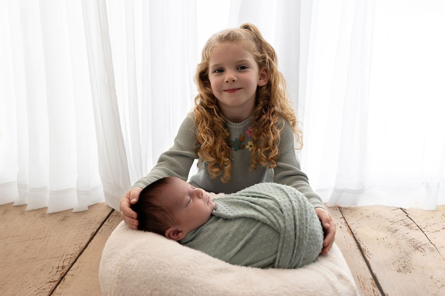 Child with curly hair sitting with baby on beige carpet by white sheer curtains.
