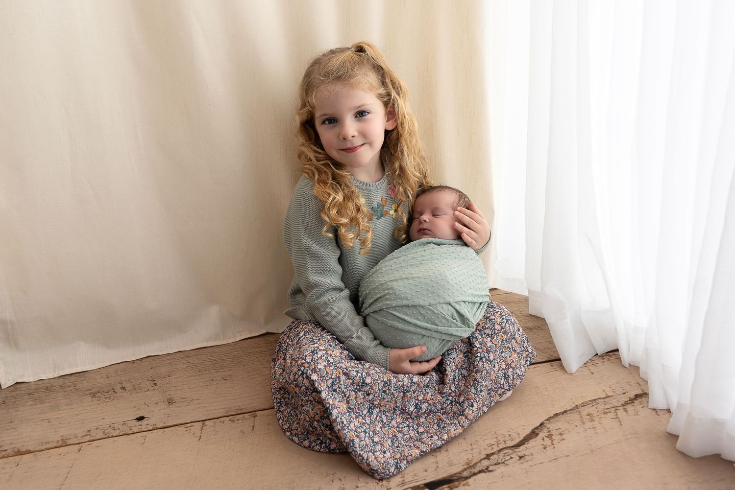 Siblings sit together against a white curtain backdrop while sharing a tender moment.