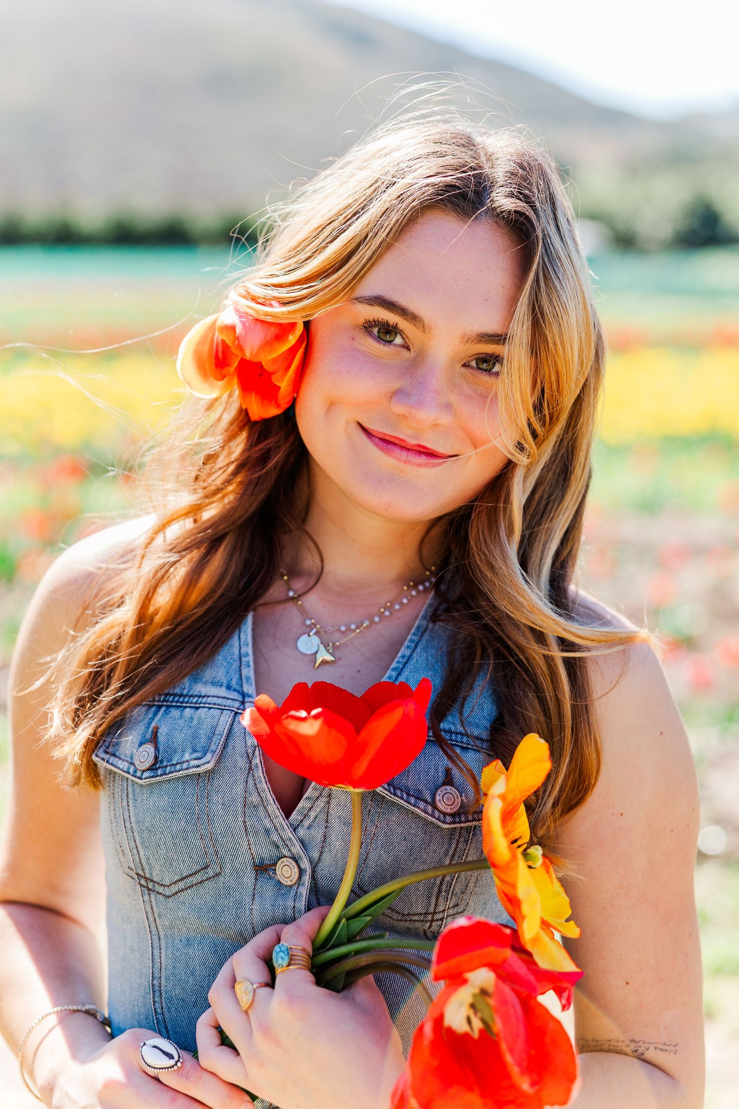 Portrait in a field of poppies wearing denim vest and holding bright red and yellow flowers on sunny day @ Underwood Family Farms