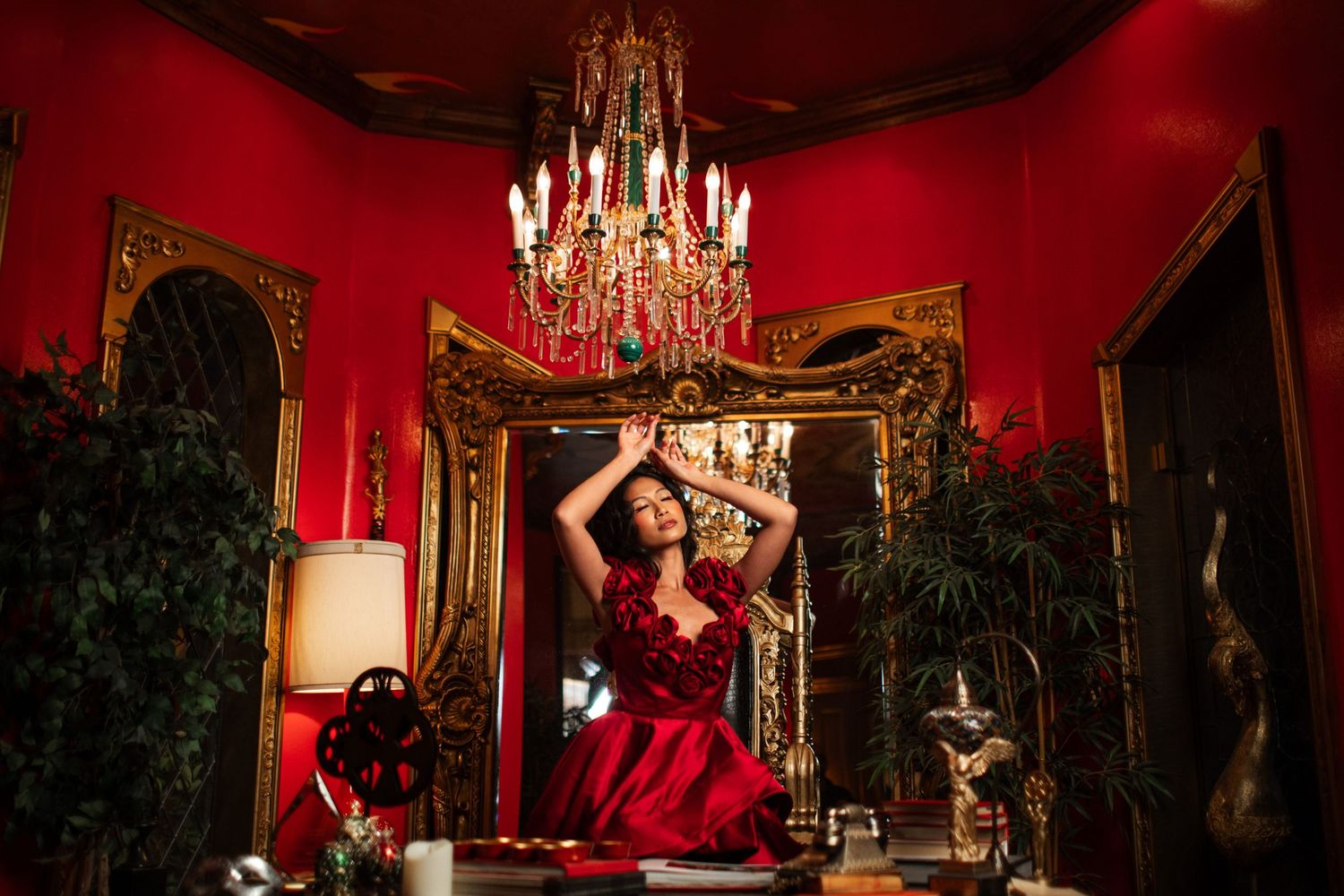 Luxurious Victorian room with red walls, ornate gold mirror, crystal chandelier and dark potted plants create dramatic ambiance.