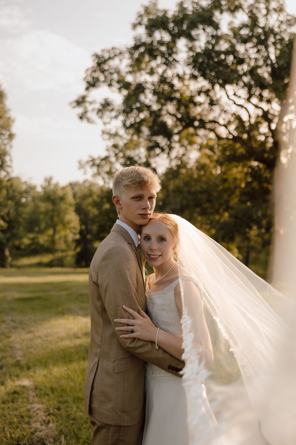 A bride and groom embrace outdoors in golden hour light, she in a flowing veil and white gown, he in a tan suit.