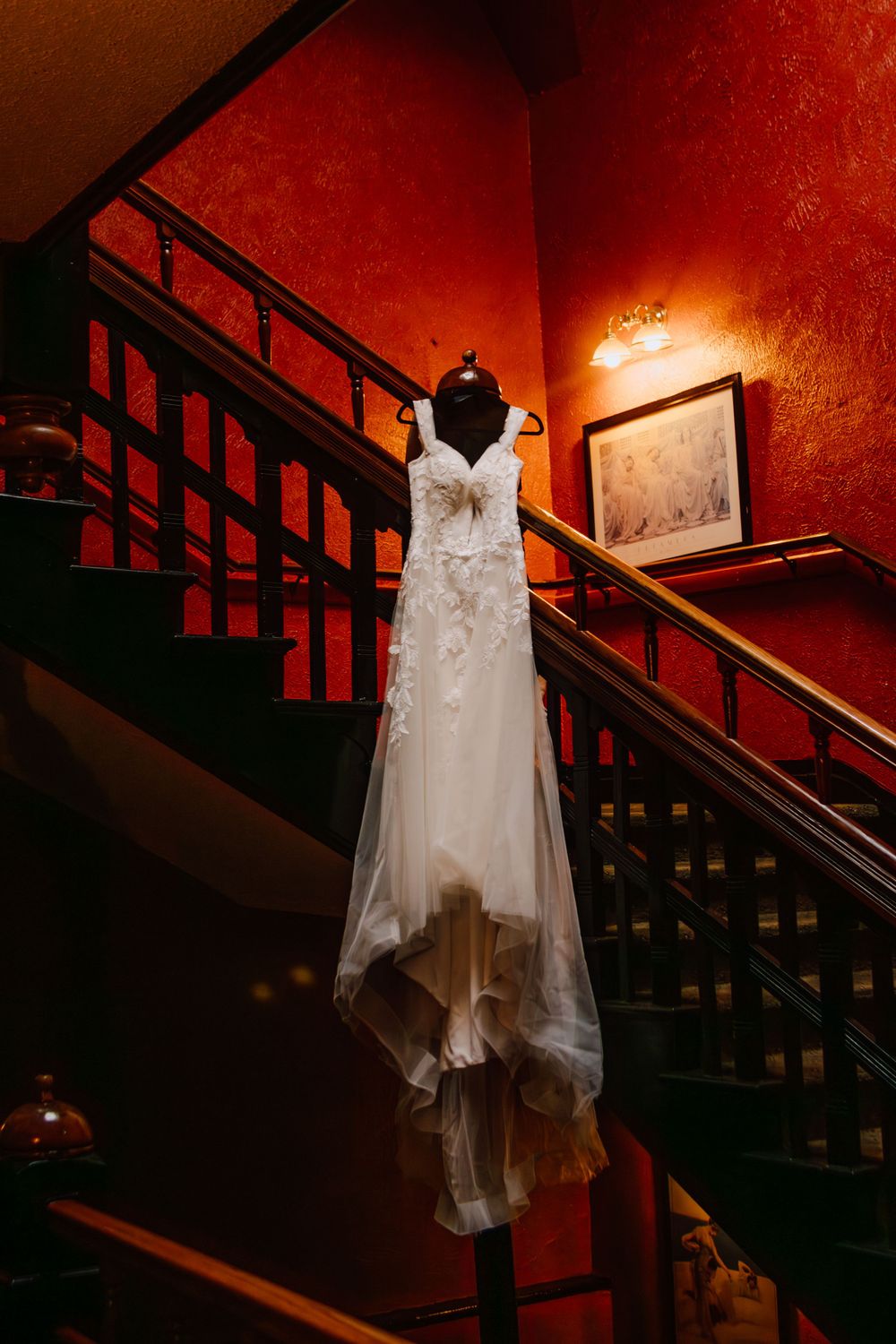 Elegant white wedding dress hanging on wooden staircase with dramatic red lighting and vintage decor.