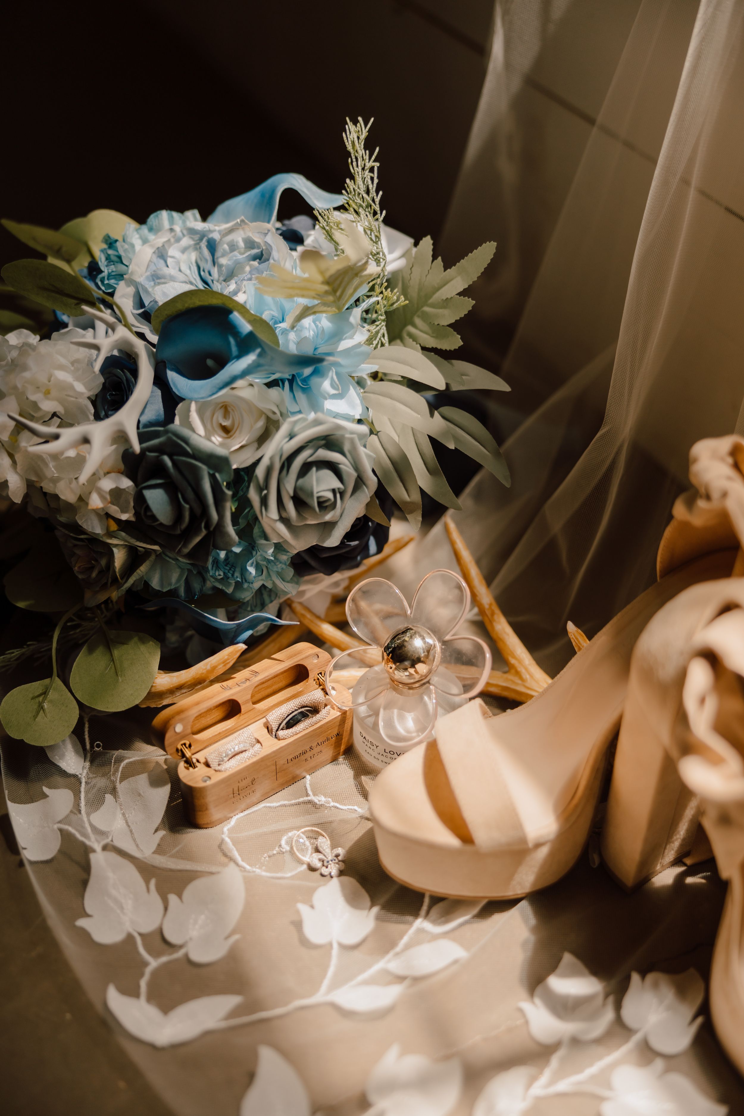 Wedding details with blue and white rose bouquet, gold rings, and blush high heel shoes on lace cloth.