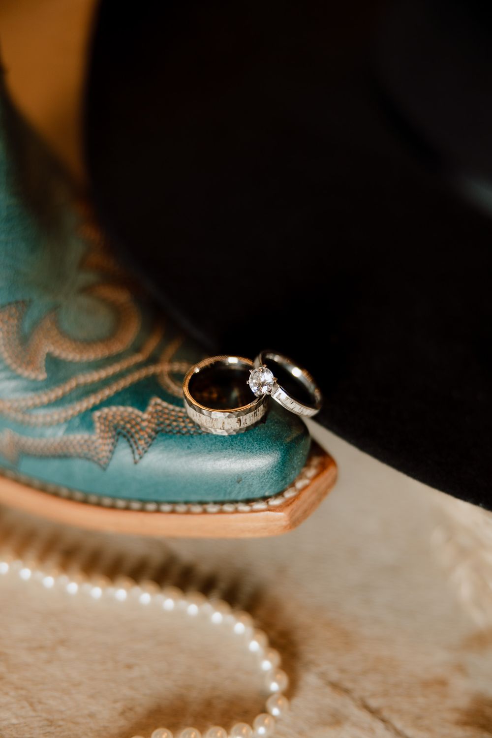 Wedding rings resting on a turquoise cowboy boot beside a black hat and pearl necklace.