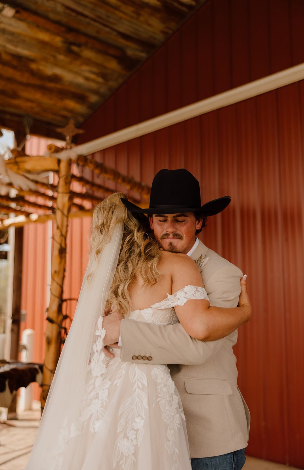 A groom in a black cowboy hat and tan suit embraces his bride in a lace wedding dress near a red barn.