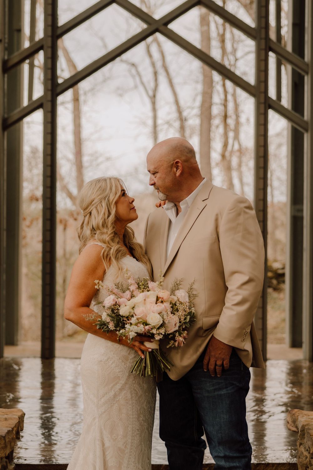 A couple shares an intimate moment in a glass chapel while holding a floral bouquet.