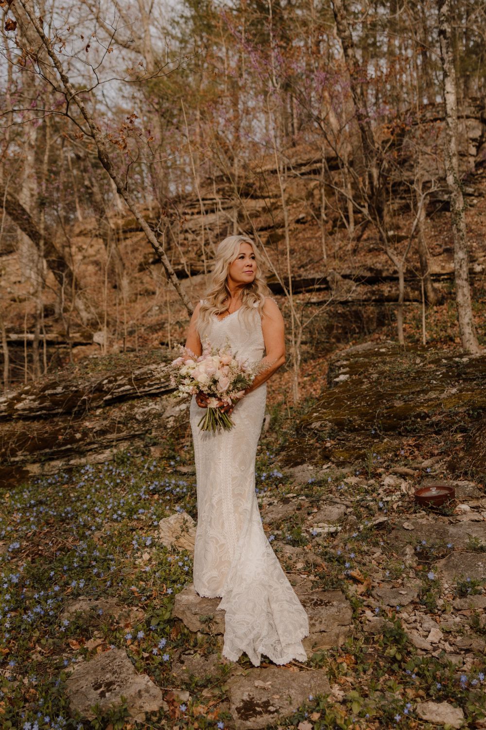 A person in a white lace wedding dress holds a bouquet against a rustic forest backdrop.