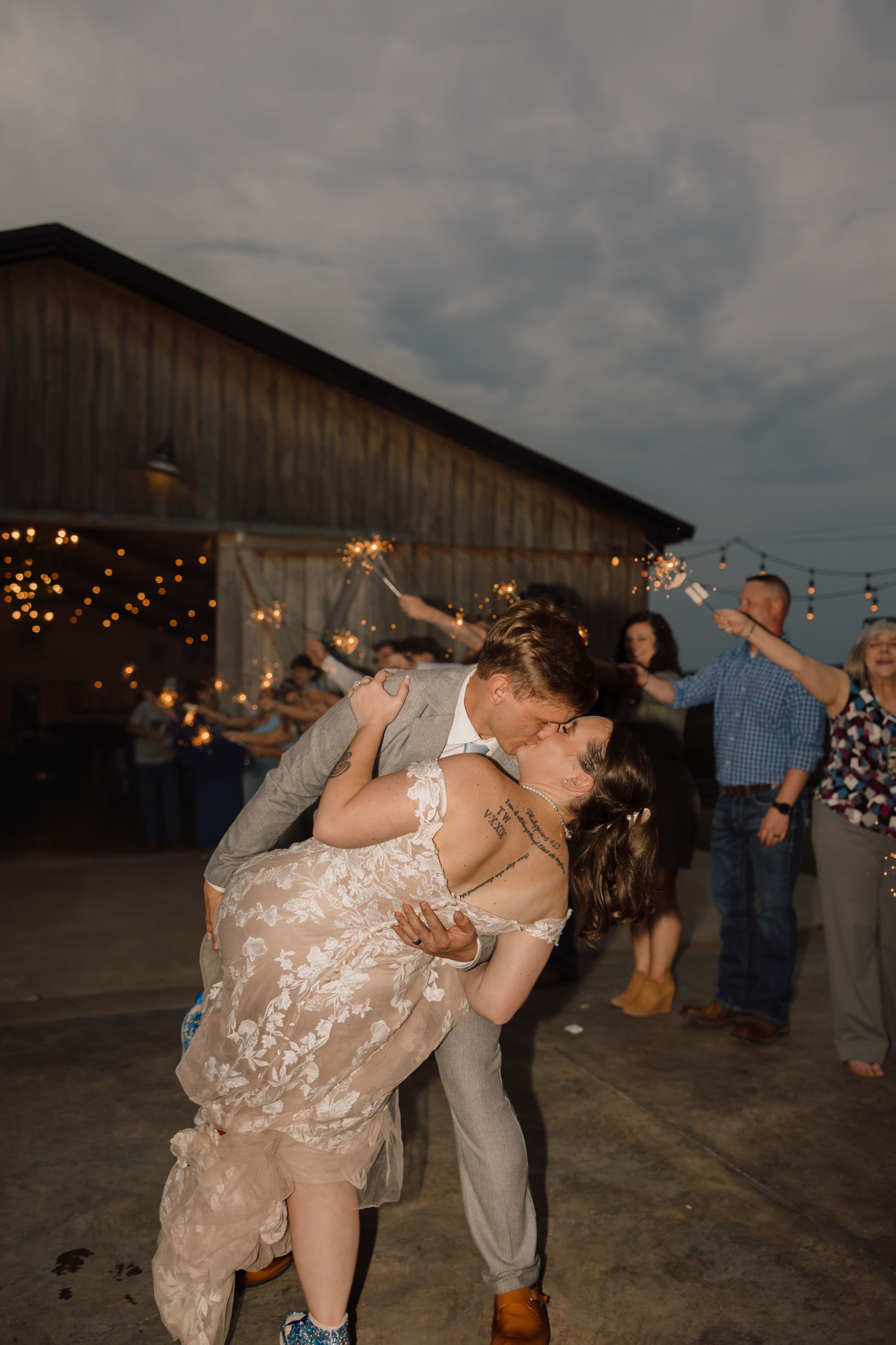 Couple shares a romantic dip kiss at rustic barn wedding reception with string lights and dark moody sky.