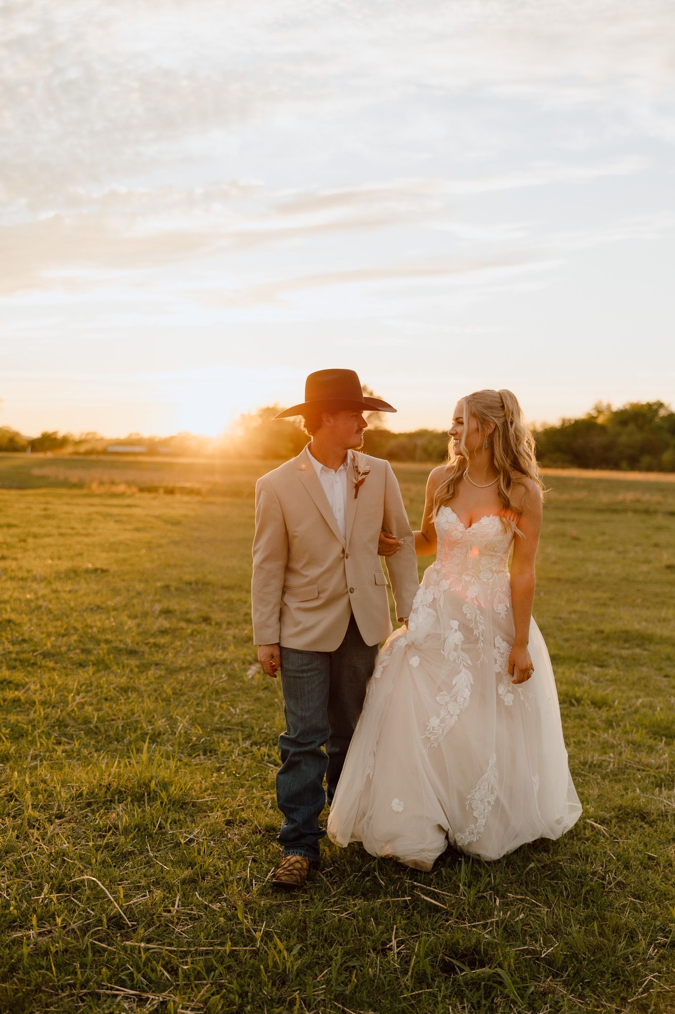 A bride and groom walk hand in hand through a green field at golden sunset, she in lace gown, he in tan suit and cowboy hat.