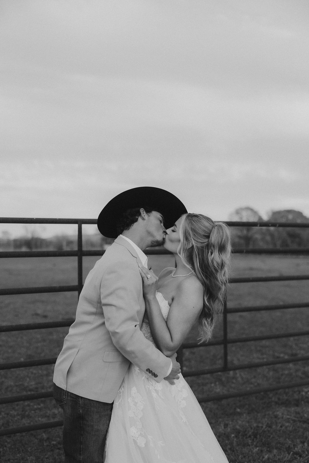 Black and white photo of a couple kissing at their rustic farm wedding, groom in cowboy hat and bride in lace gown.